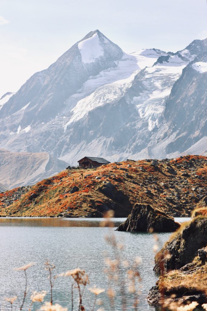 Lac entouré d'herbe orangée et cabane en bois, des sommets de montagne en arrière plan